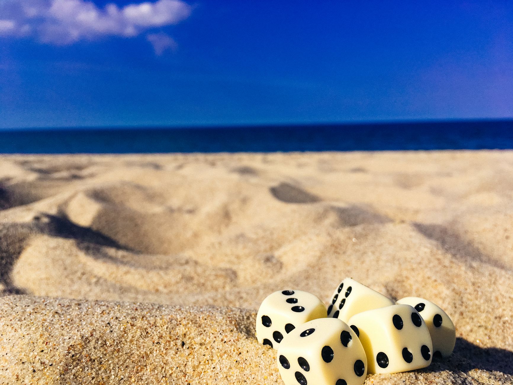 Five dice resting on a sunlit beach with ocean in the background, perfect for summer and leisure themes.