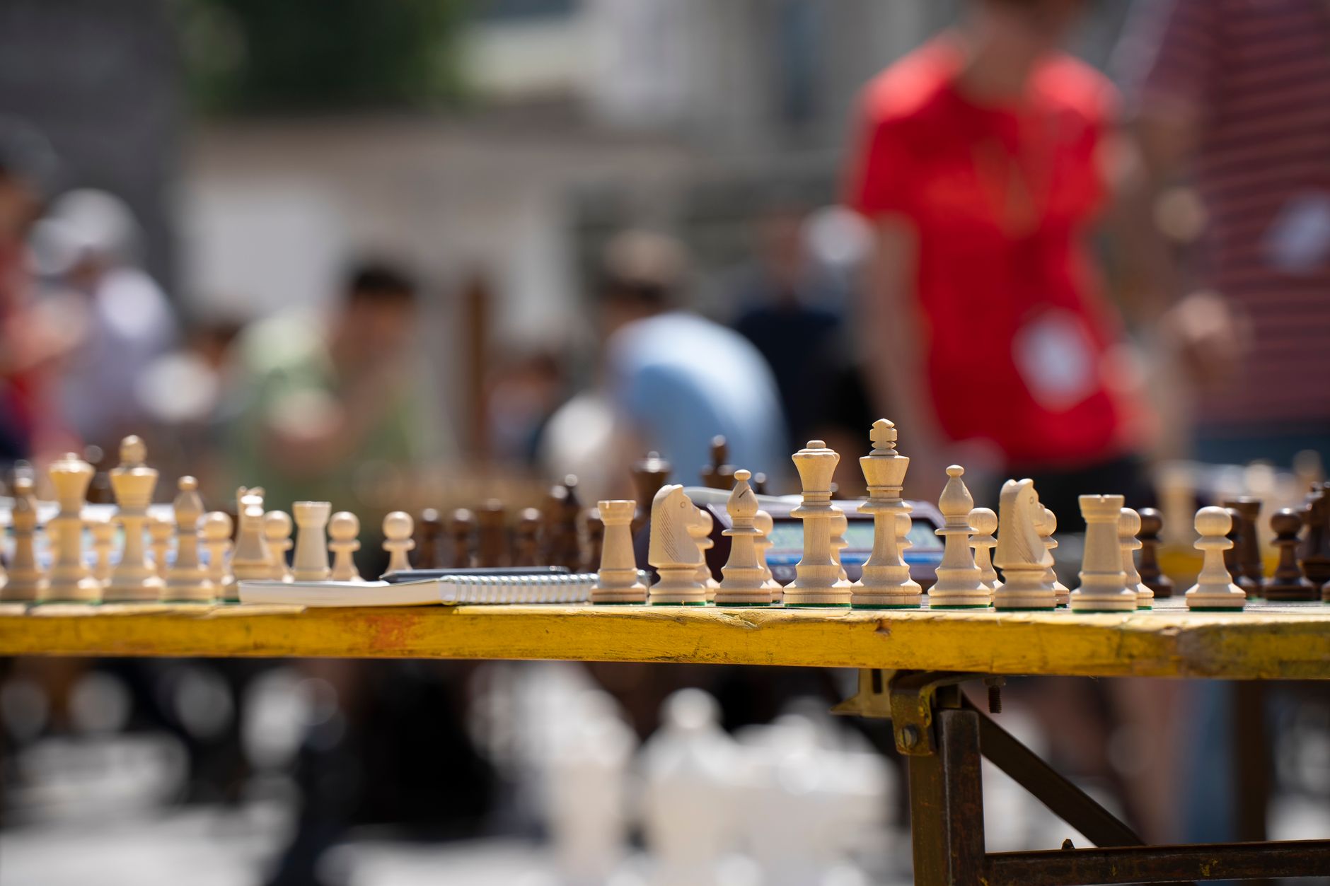 Chess pieces on a table with blurred people in the background, capturing a vibrant outdoor scene.