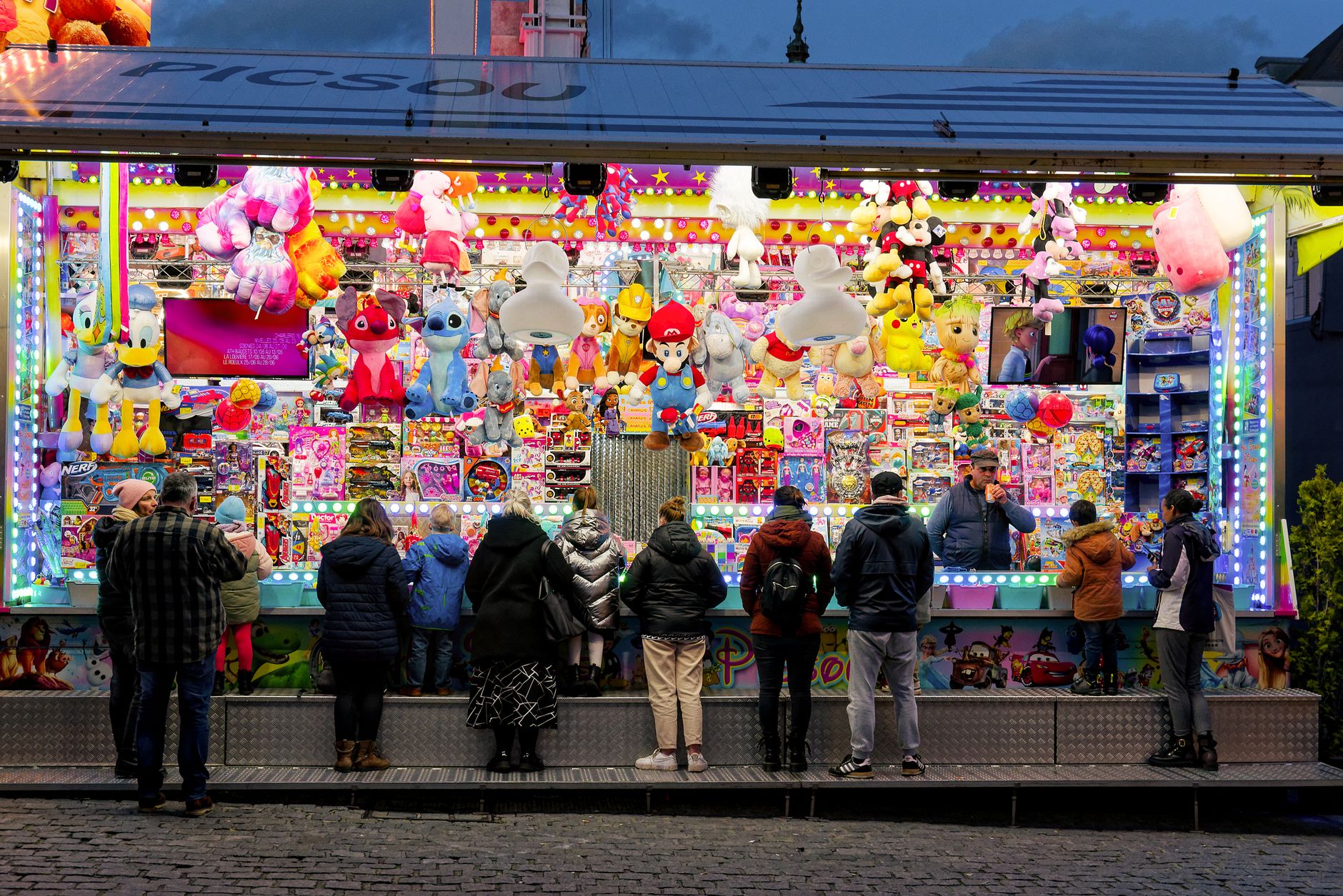 People at an illuminated carnival game stand with plush toys and vibrant lights at night.