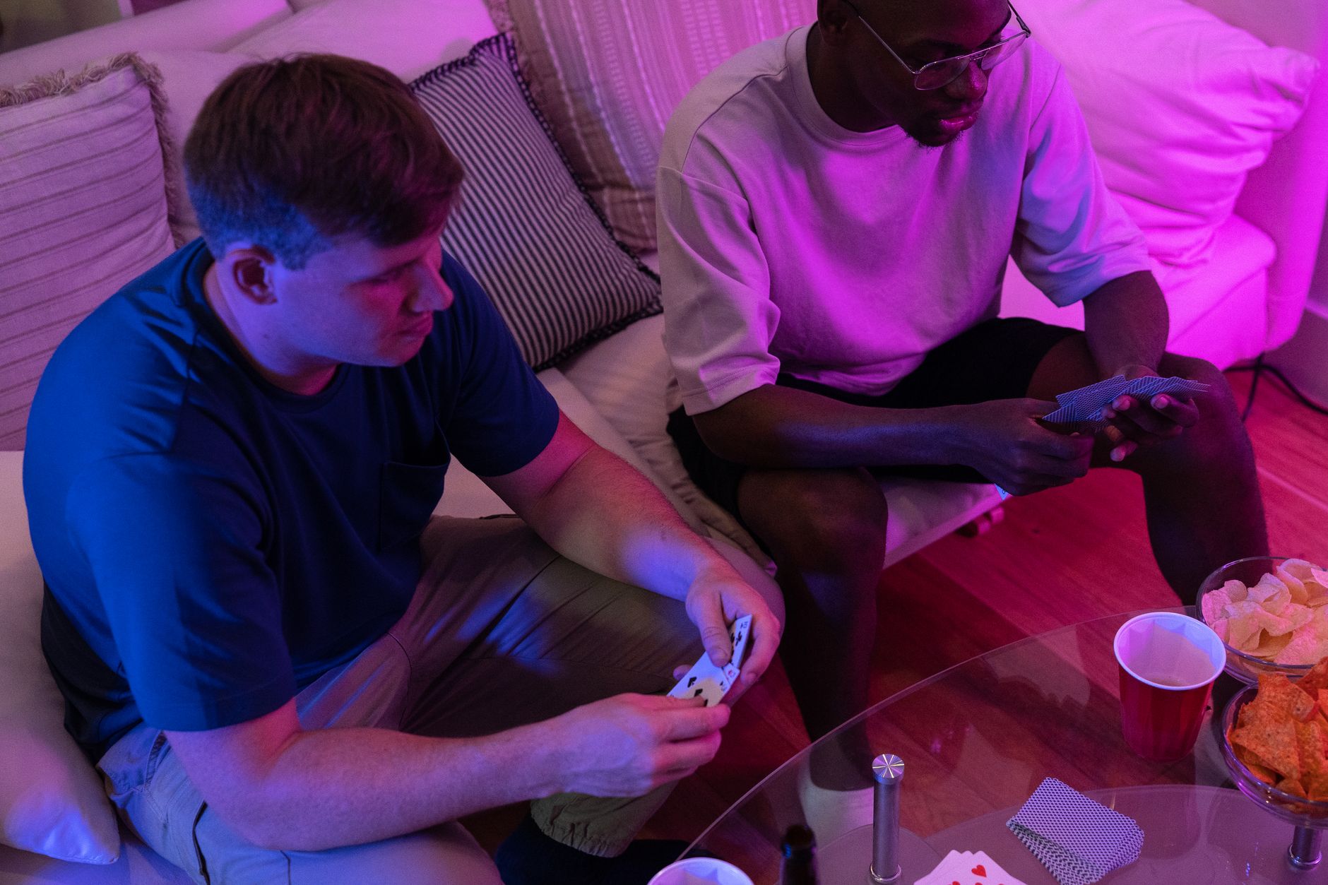 Two friends enjoy a casual card game at home under warm pink lighting.