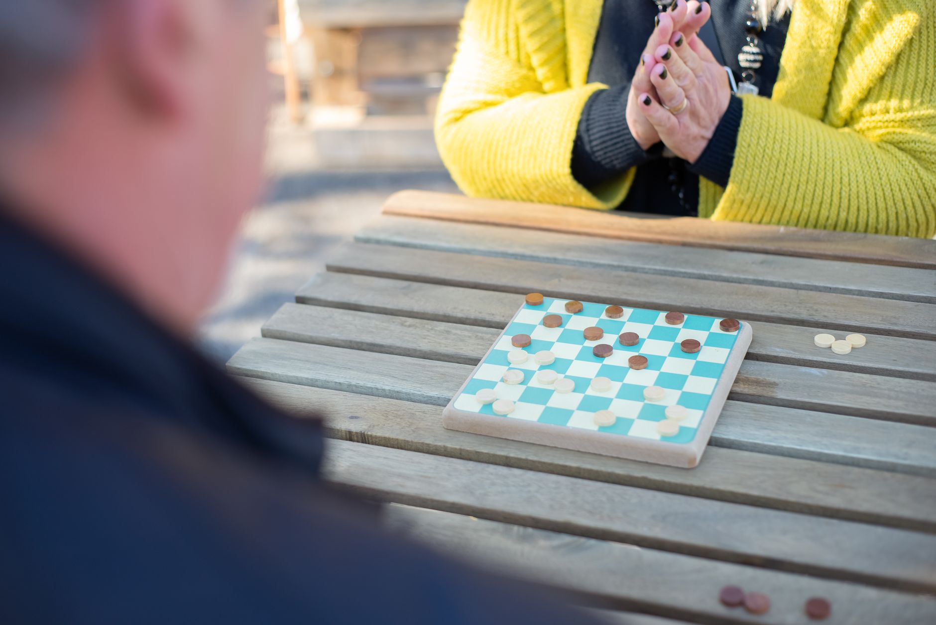 Adults enjoying a strategic board game on a wooden table outdoors in bright sunlight.