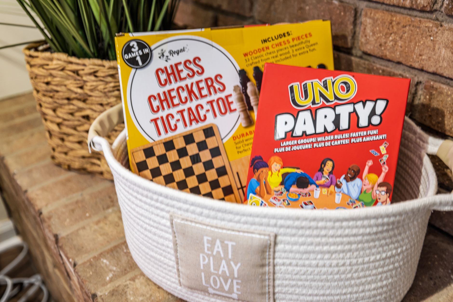 Basket with board games including Chess, Checkers, Tic-Tac-Toe, and UNO Party on a brick fireplace.