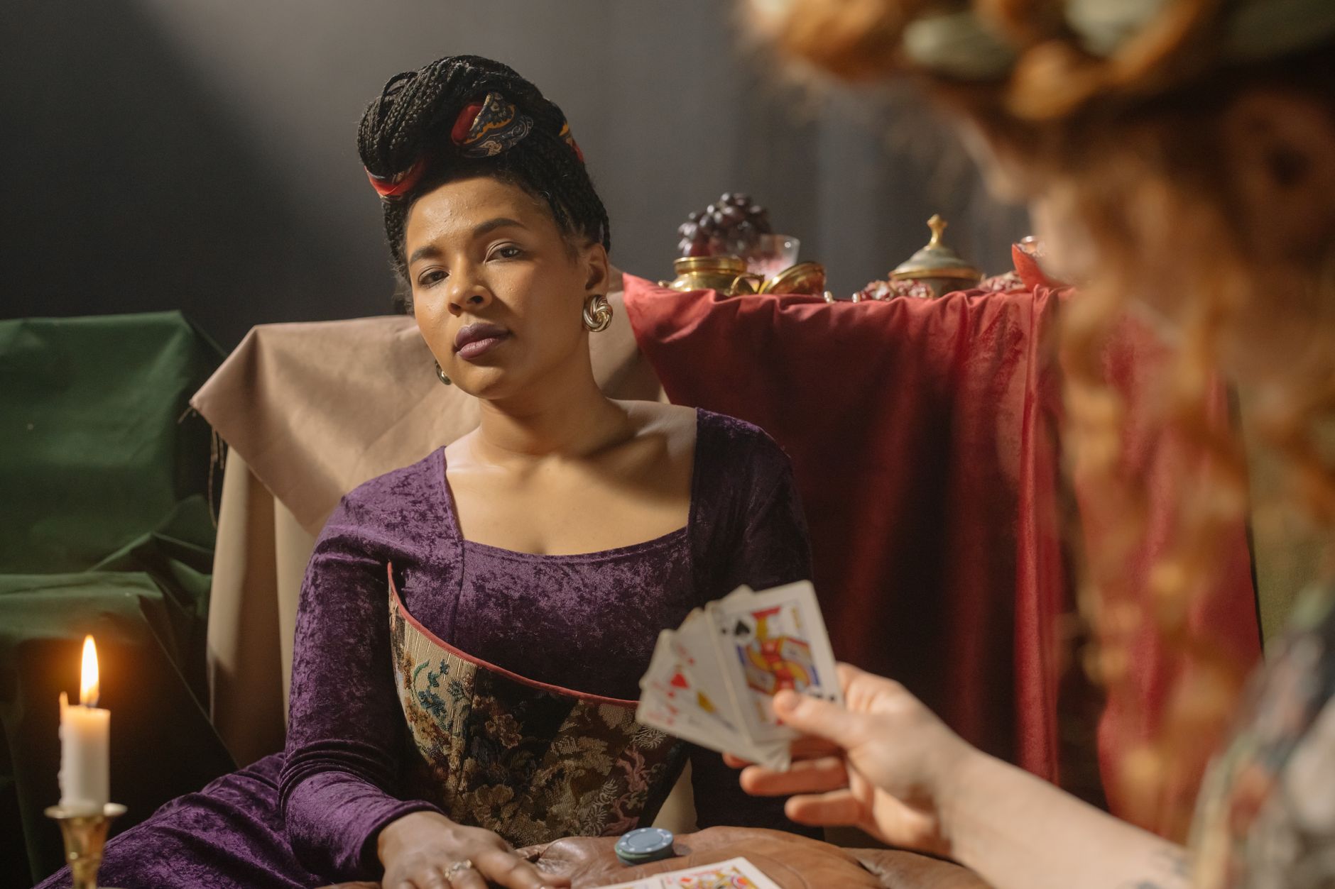 A woman in period costume gazing while holding playing cards in a candlelit room.