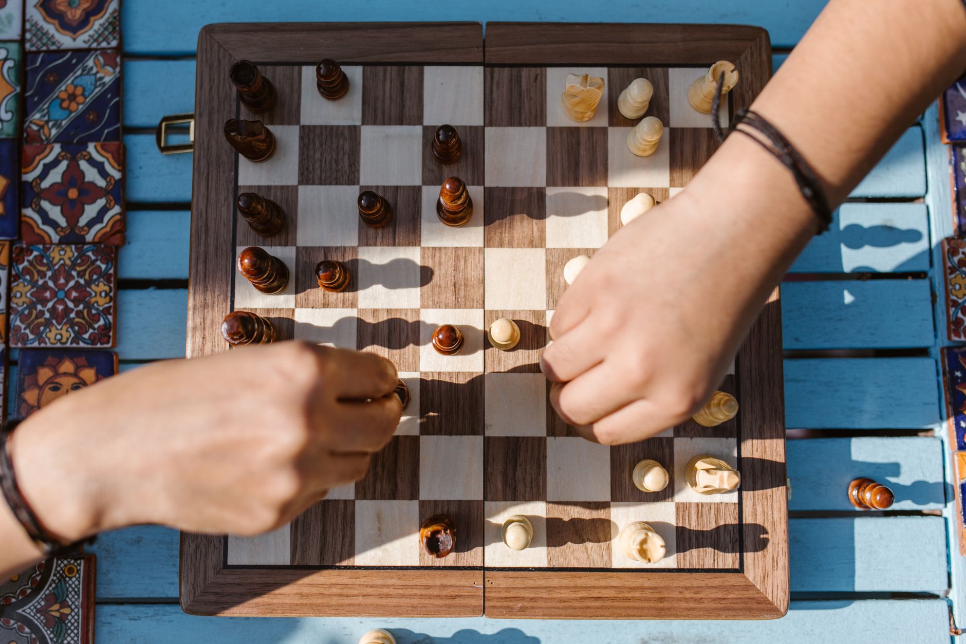 Close-up of hands playing chess on a wooden board outdoors under bright sunlight.