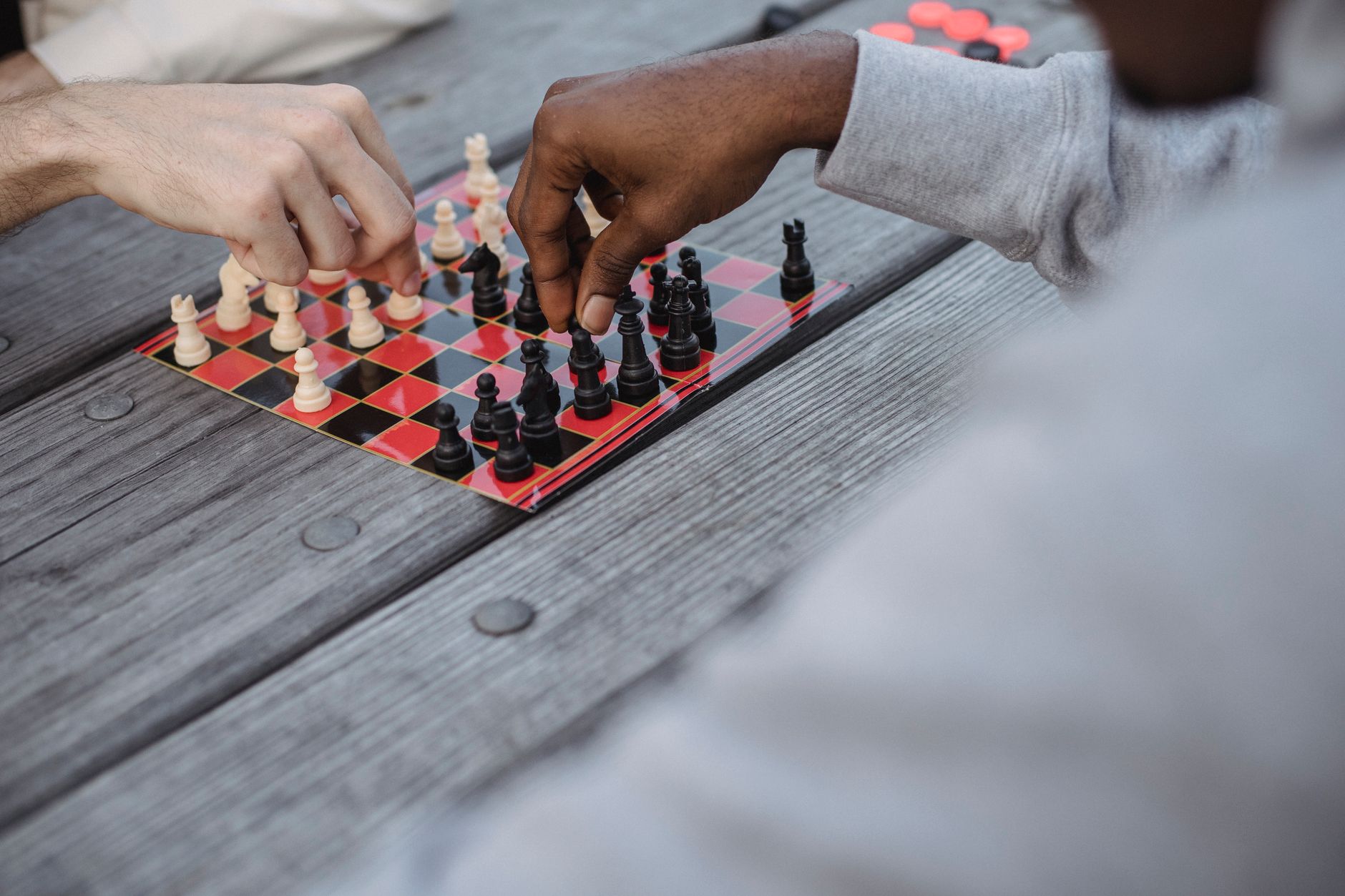High angle of crop faceless young diverse male friends playing chess sitting at wooden table in daylight